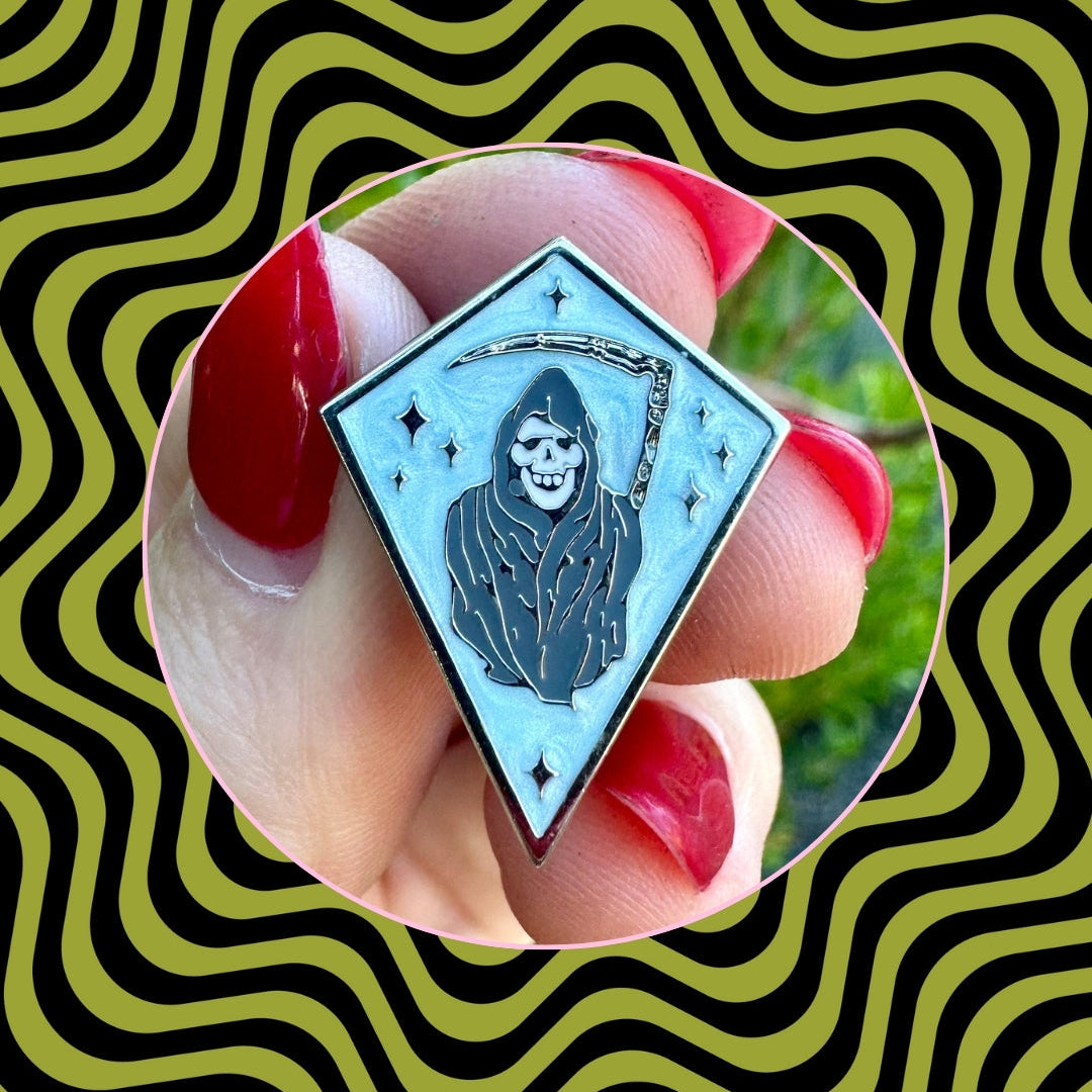 Diamond-shaped enamel pin with grim reaper design held by a hand against a wavy patterned background.