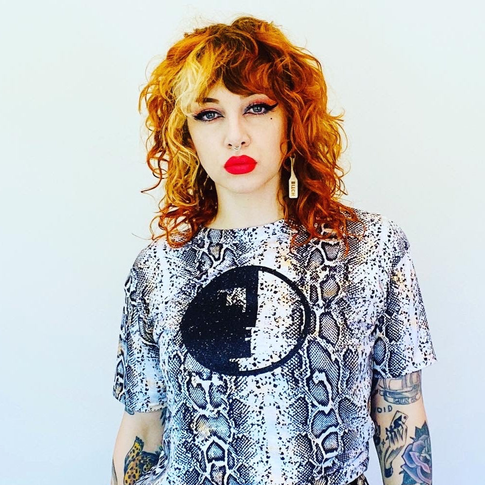 Person with red hair and tattoos wearing a patterned top and black pants against a light blue background