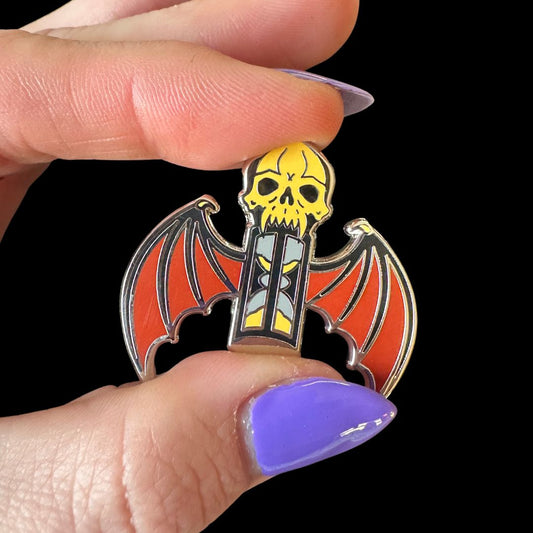 Enamel pin with bat and skull design held between fingers against a black background