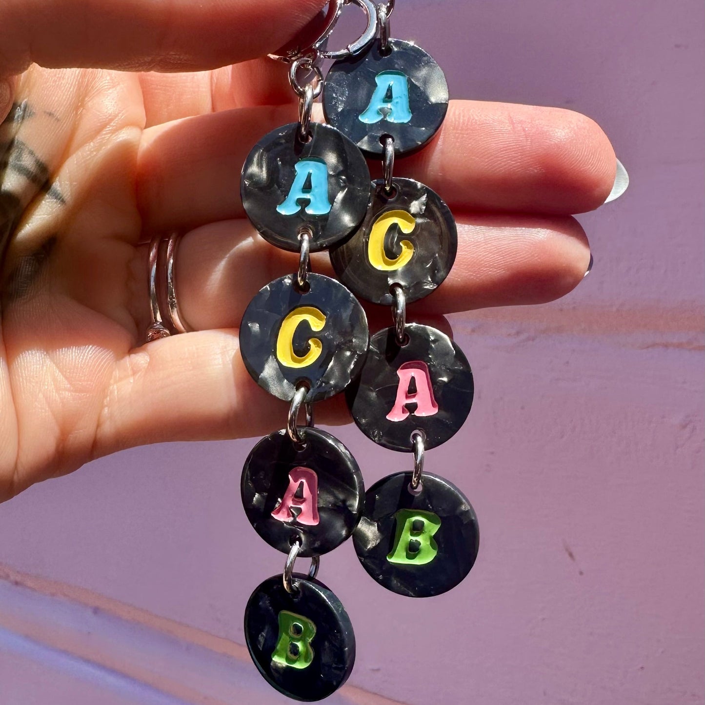 A close-up of a hand holding a pair of dangling earrings made of round black marbled discs, each printed with a colorful letter (A, C, A, B) on a pink wall background.