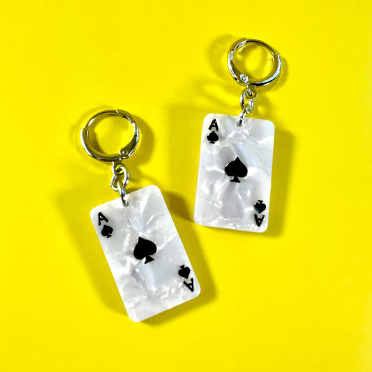 A pair of white acrylic earrings designed to look like Ace of Spades playing cards, featuring silver-tone key rings at the top, displayed on a bright yellow background.