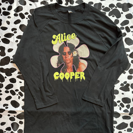 A black long-sleeve graphic t-shirt featuring a large front print of Alice Cooper with yellow text and a flower graphic, laid flat on a black and white cow-print fabric background.