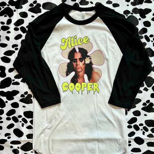 A 3/4-sleeve raglan baseball t-shirt featuring a black body and black sleeves, with a white front graphic of Alice Cooper inside a flower with yellow text. The shirt is laid flat on a black and white cow-print background.