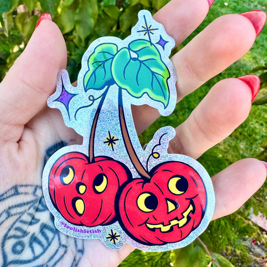 Hand with tattoo holding sparkly sticker of two retro jack o lantern pumpkin faces on a cherry style stem