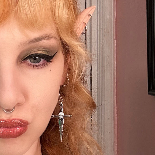 Person with red hair and elf ears in a room with framed pictures on the wall with a punk goth metal enamel skull dagger earring.