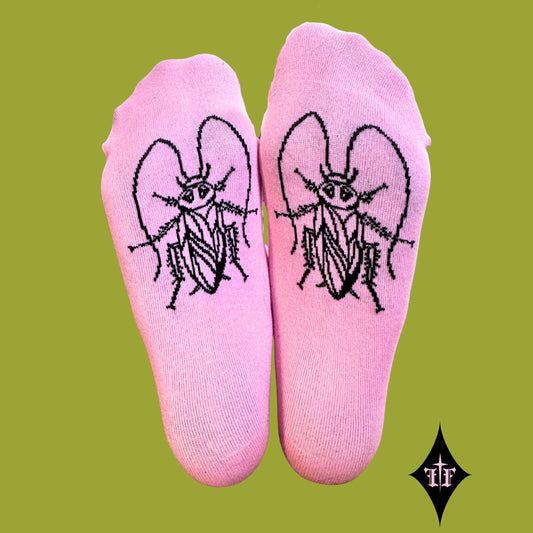 A pair of light pink crew socks with a black embroidered winged insect design on the top of each foot, displayed against a solid olive green background.