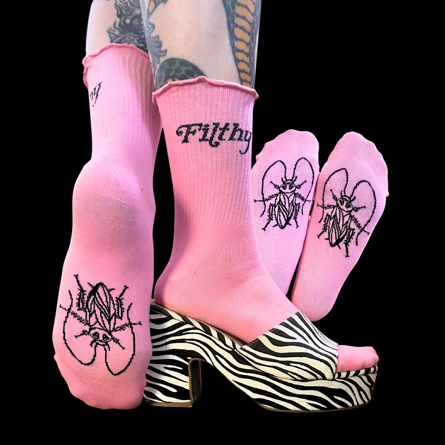 Pink crew socks with black embroidered gothic-style text and insect illustrations, shown on a model's feet paired with zebra-print platform heels against a black background.
