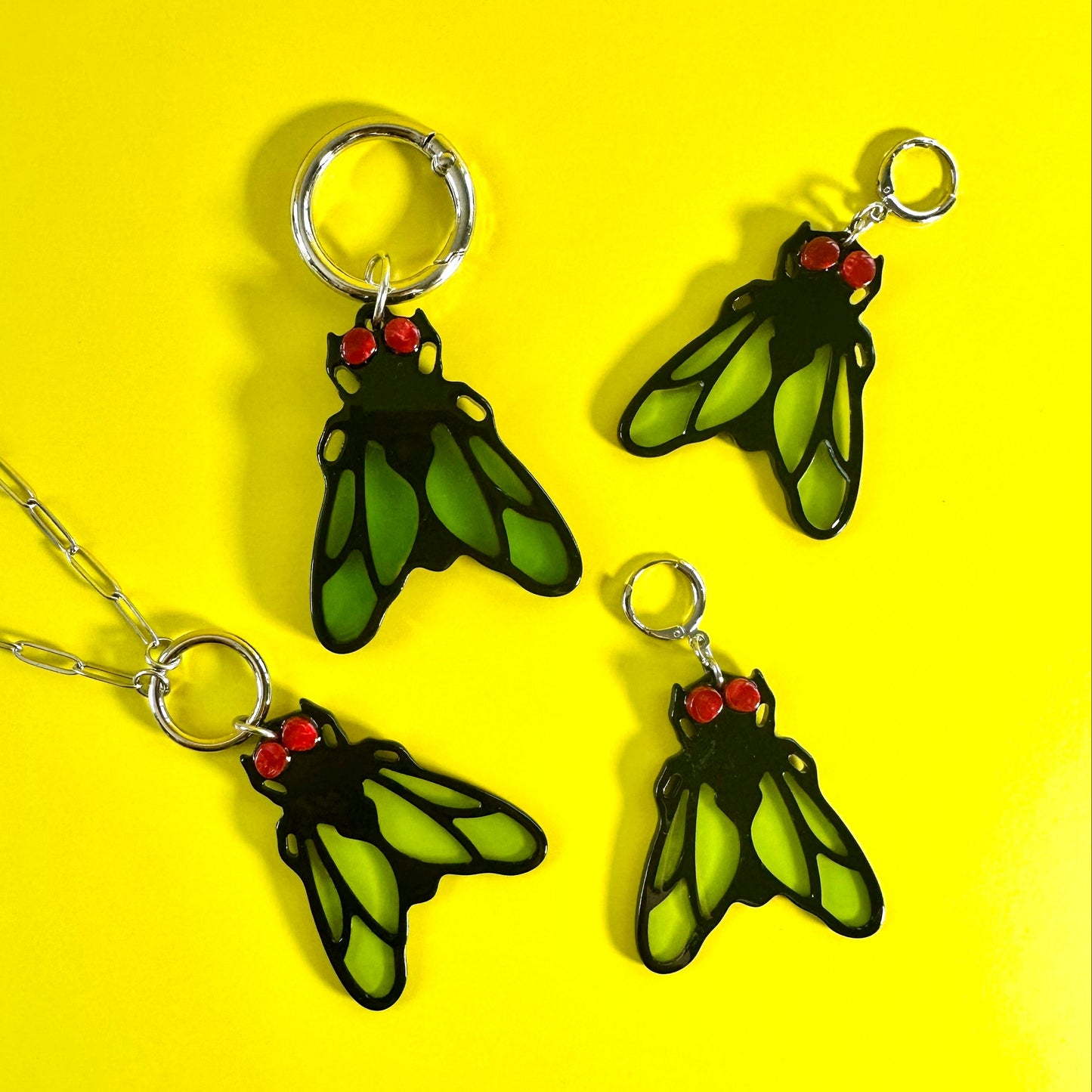 Three insect-themed jewelry charms or pendants with silver key rings and chain, featuring green wings with black outlines and red heads, arranged on a bright yellow background.