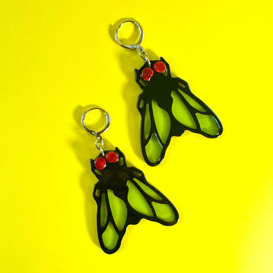 A pair of dangling earrings shaped like cicadas, featuring bright green wings outlined in black with small red eyes and silver-tone hooks, displayed on a solid yellow background.