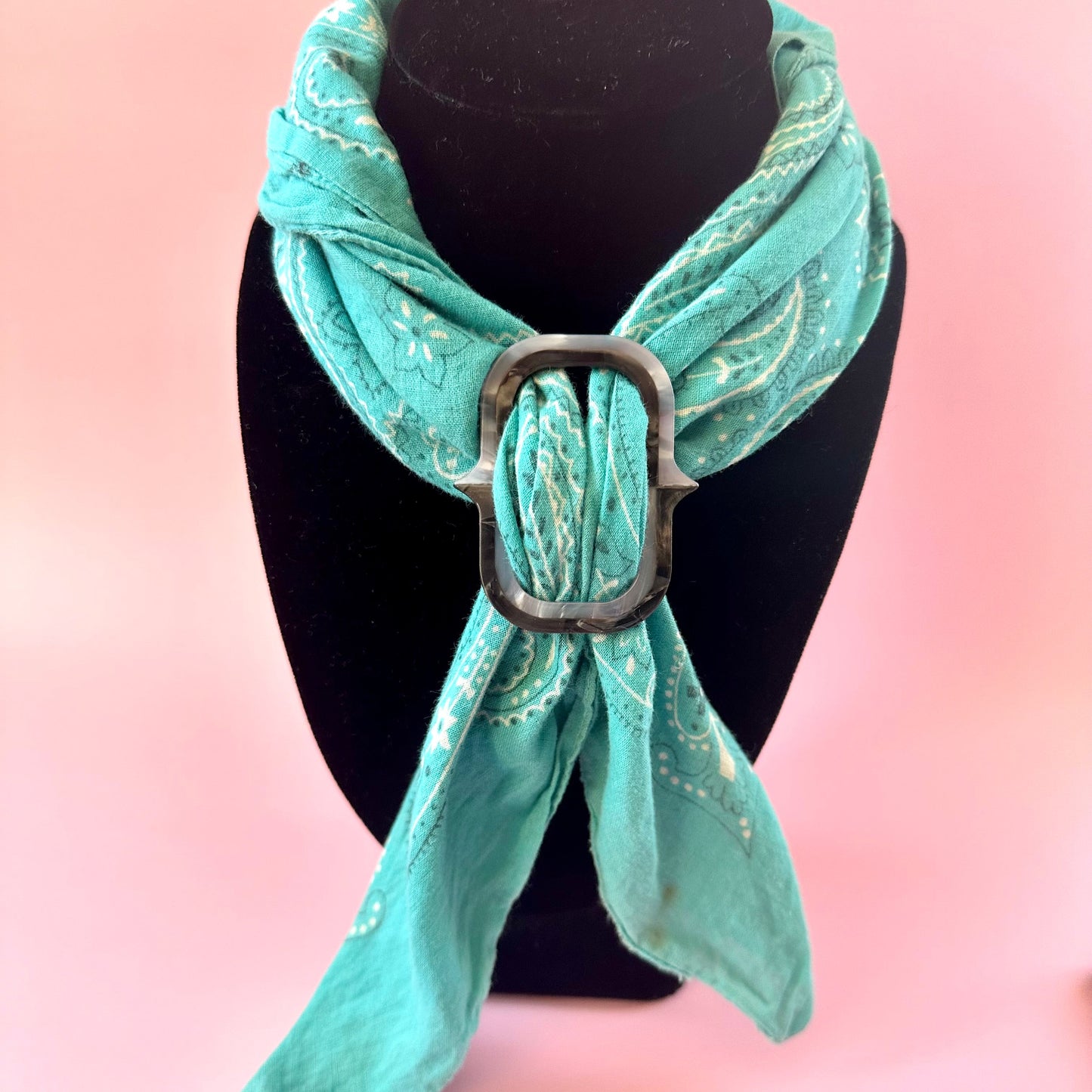 A mint green paisley bandana scarf with a large silver metal buckle, displayed on a black mannequin bust against a soft pink background.