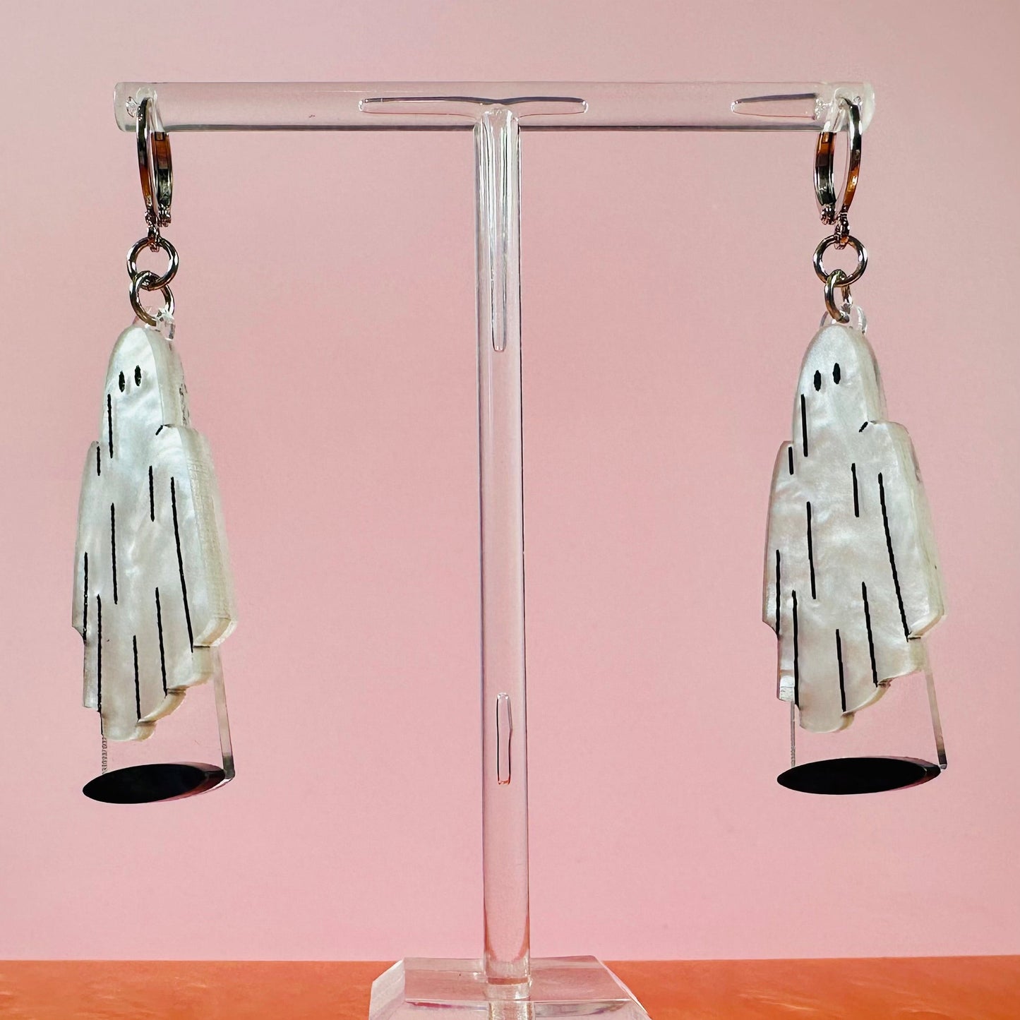 Pair of ghost-shaped earrings on a clear stand with a pink background