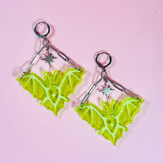 Pair of green bat-shaped earrings on a pink background