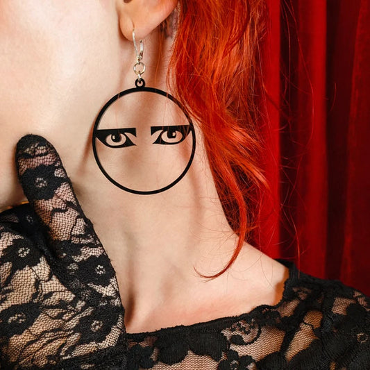 Person wearing a black lace garment and eye-shaped earrings with a red curtain background