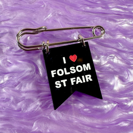 Black flag-shaped keychain with 'I ❤️ FOLSOM ST FAIR' text on a purple textured background