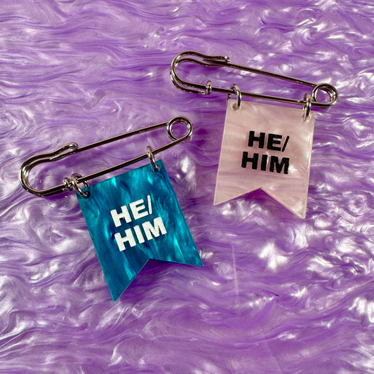 Two safety pins with 'HE/HIM' labels on a purple marbled background