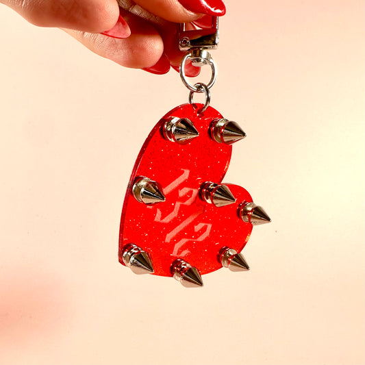Red heart-shaped keychain with spikes held by a hand against a beige background