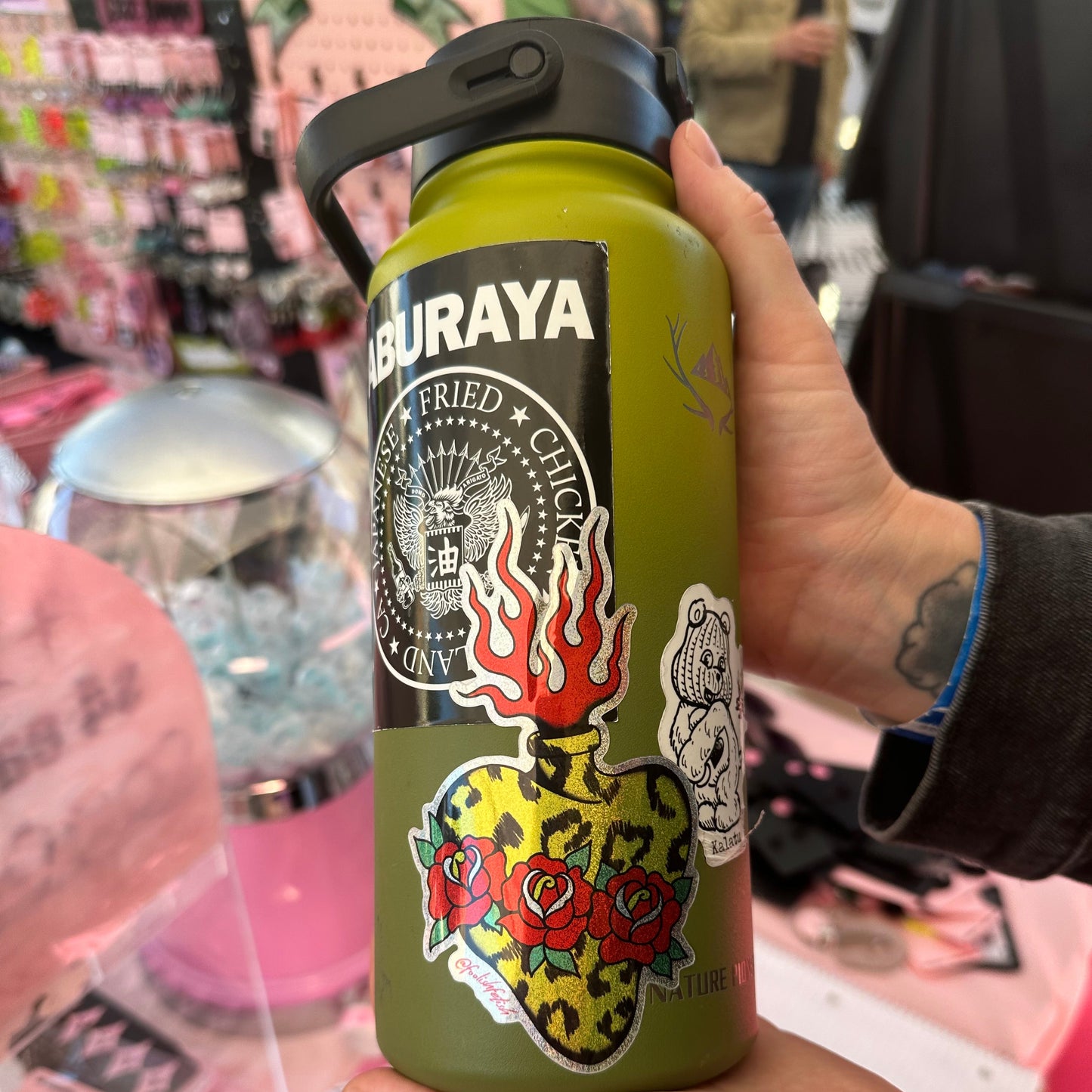 A person holds a green insulated water bottle with a black lid and multiple colorful stickers, including a skull, flames, and roses. The background is a busy retail display with pink items.