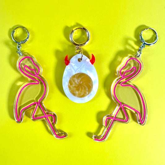 Three flamingo-themed earrings arranged on a bright yellow background. Two earrings feature pink flamingo outlines with silver-tone rings, and the center keychain features a white deviled egg pendant with small red horns and a gold swirl center.