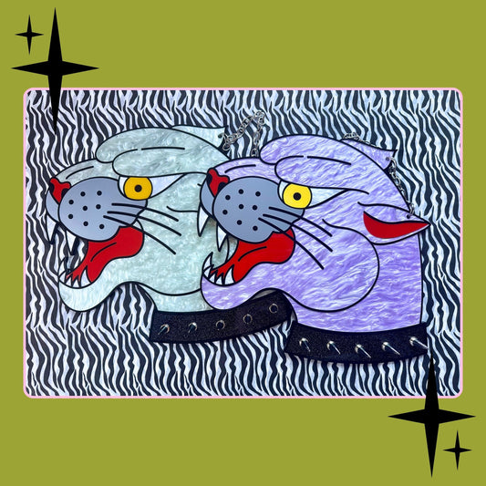 A stylized art print featuring two cat heads in profile, one pale blue-gray and one lavender-purple, both with red tongues, yellow eyes, black spots, and black studded collars, set against a black-and-white zebra pattern. The print is centered on an olive green background with black starburst accents in the corners.
