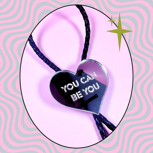 Heart-shaped pendant with 'You Can Be You' text on a pink and gray wavy background