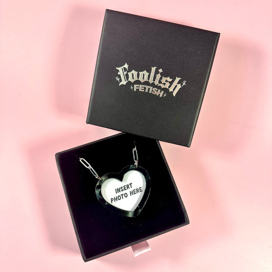 Heart-shaped pendant in a black box with 'Foolish Fetish' branding on a pink background