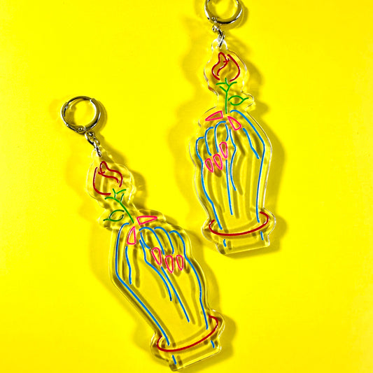 A pair of acrylic earrings shaped like hands holding flowers in a vase, displayed on a bright yellow background.