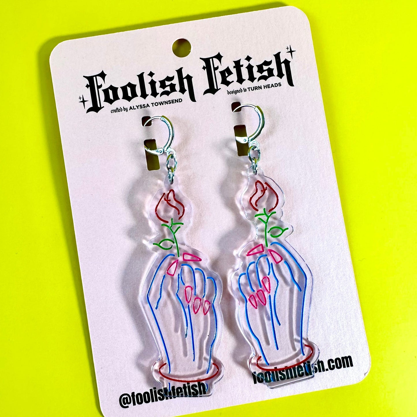 A pair of dangling earrings on a white retail card. The earrings are made of clear acrylic with a printed neon-style design of skeletal hands holding pink roses with green stems.