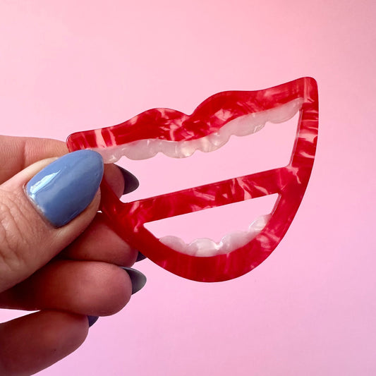 A hand with blue nail polish holds a red and white marbled lip-shaped scarf ring against a solid pink background.