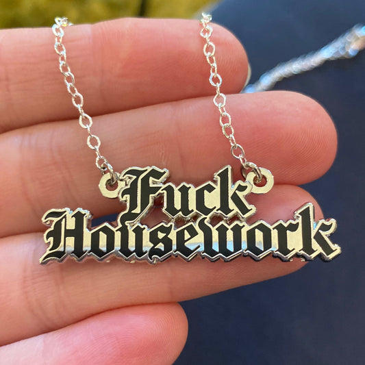 A silver-colored enamel pendant necklace with the text 'FUCK HOUSEWORK' displayed, held in a person's hand.