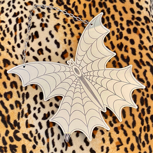 A silver-colored mini spiderweb butterfly wall hanging with a chain for mounting, laid out on a leopard print fabric.