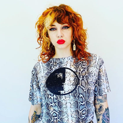 Person with red hair and tattoos wearing a patterned top and black pants against a light blue background