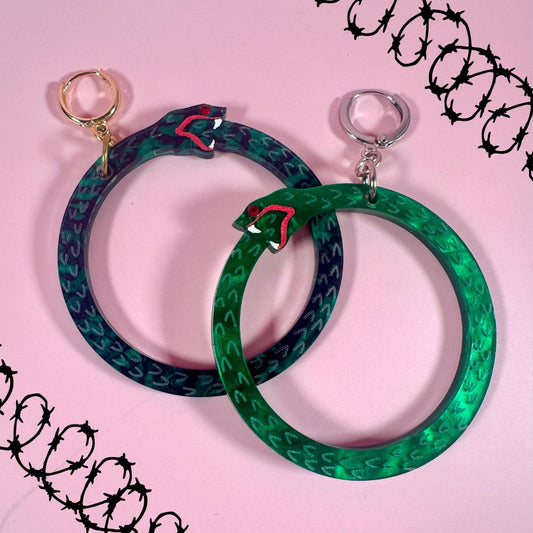 Two green and blue patterned hoop earrings on a pink background with black barbed wire design.