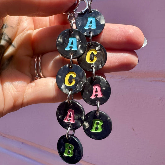 A close-up of a hand holding a pair of dangling earrings made of round black marbled discs, each printed with a colorful letter (A, C, A, B) on a pink wall background.