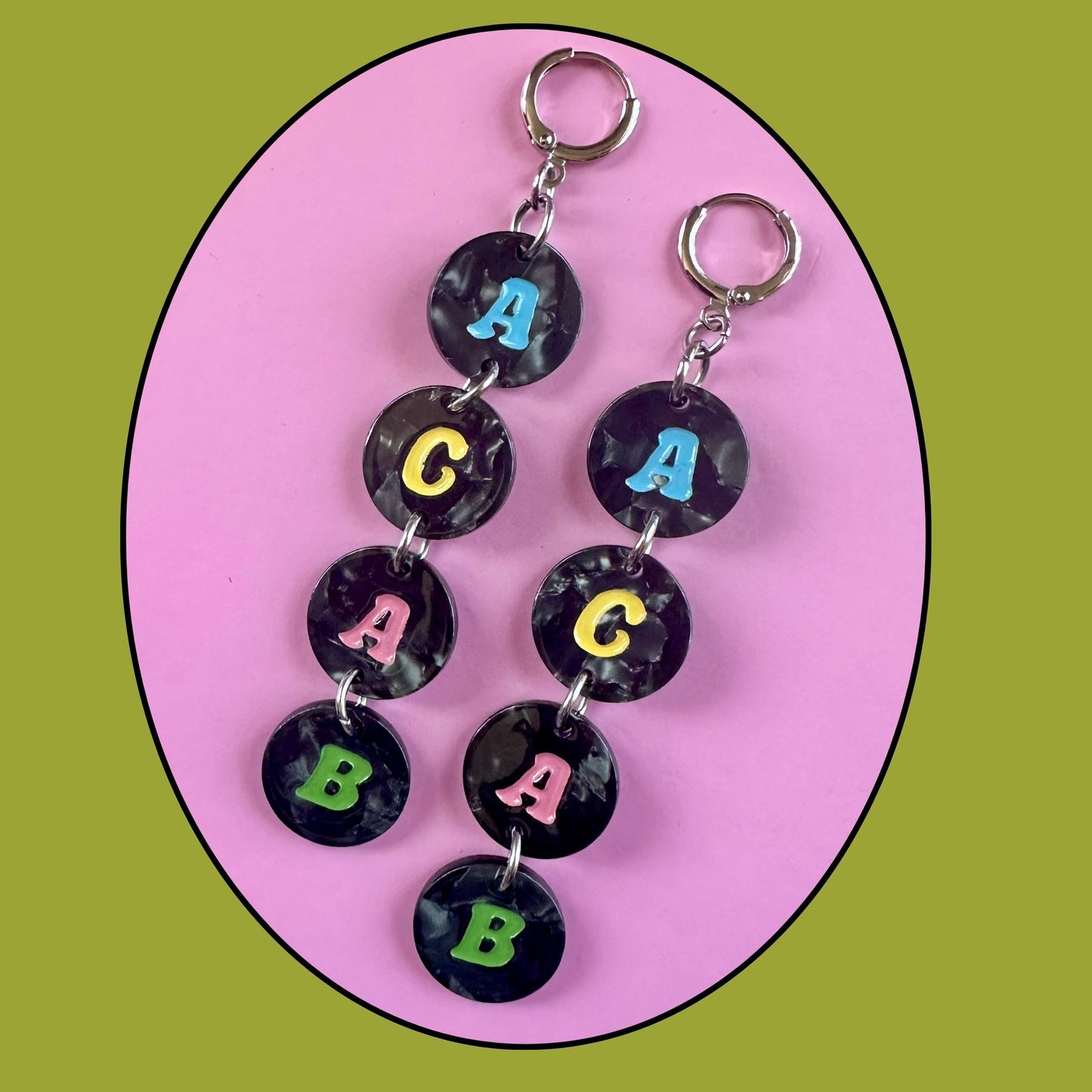 Two matching earrings with dangling round black marbled beads printed with colorful letters A, C, A, and B, displayed on a pink oval background with an olive green outer backdrop.