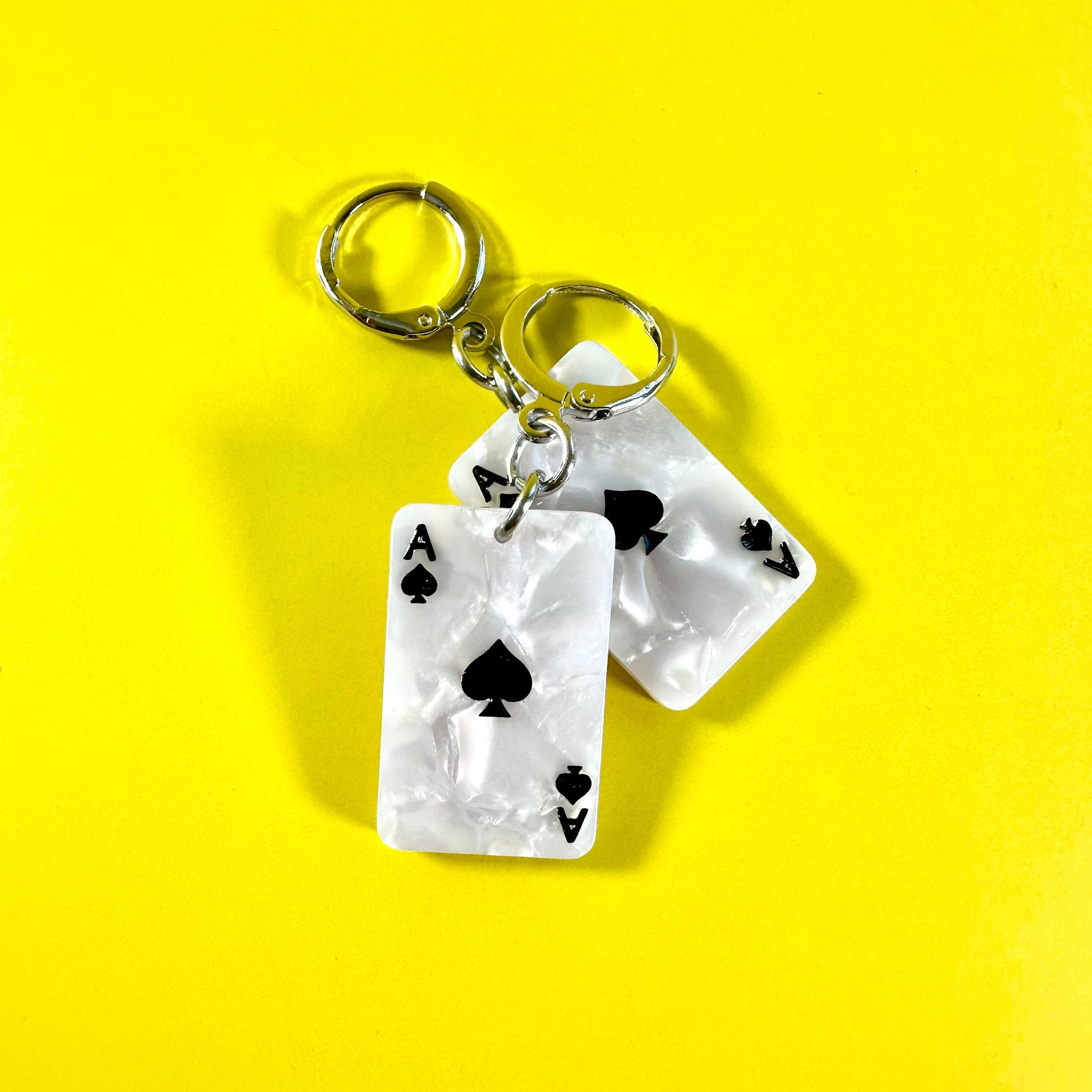Two acrylic earrings shaped like Ace of Spades playing cards with silver split rings, displayed on a bright yellow background.