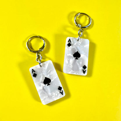 A pair of white acrylic earrings designed to look like Ace of Spades playing cards, featuring silver-tone key rings at the top, displayed on a bright yellow background.