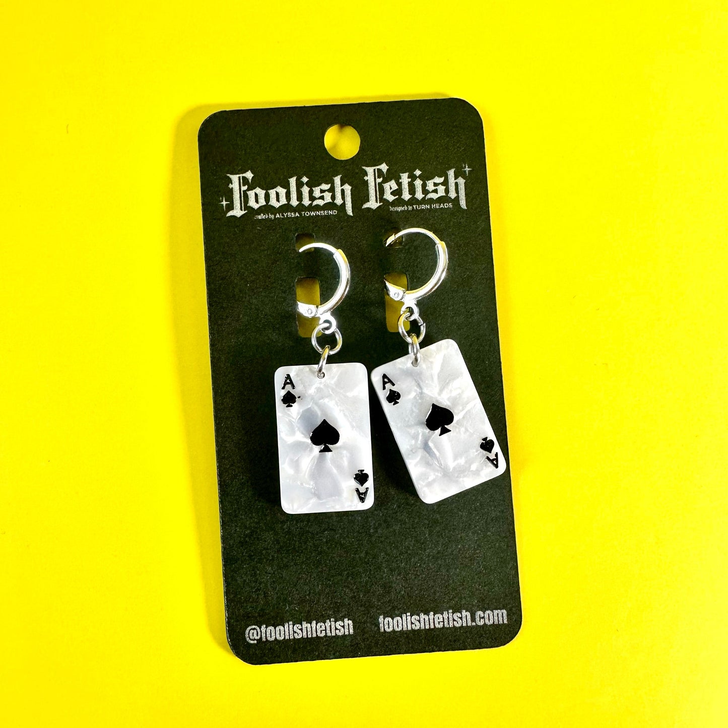 A pair of silver-toned dangling earrings shaped like playing cards, mounted on a black display card with the brand name 'Foolish Fetish' printed at the top, set against a bright yellow background.