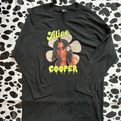 A black long-sleeve graphic t-shirt featuring a large front print of Alice Cooper with yellow text and a flower graphic, laid flat on a black and white cow-print fabric background.