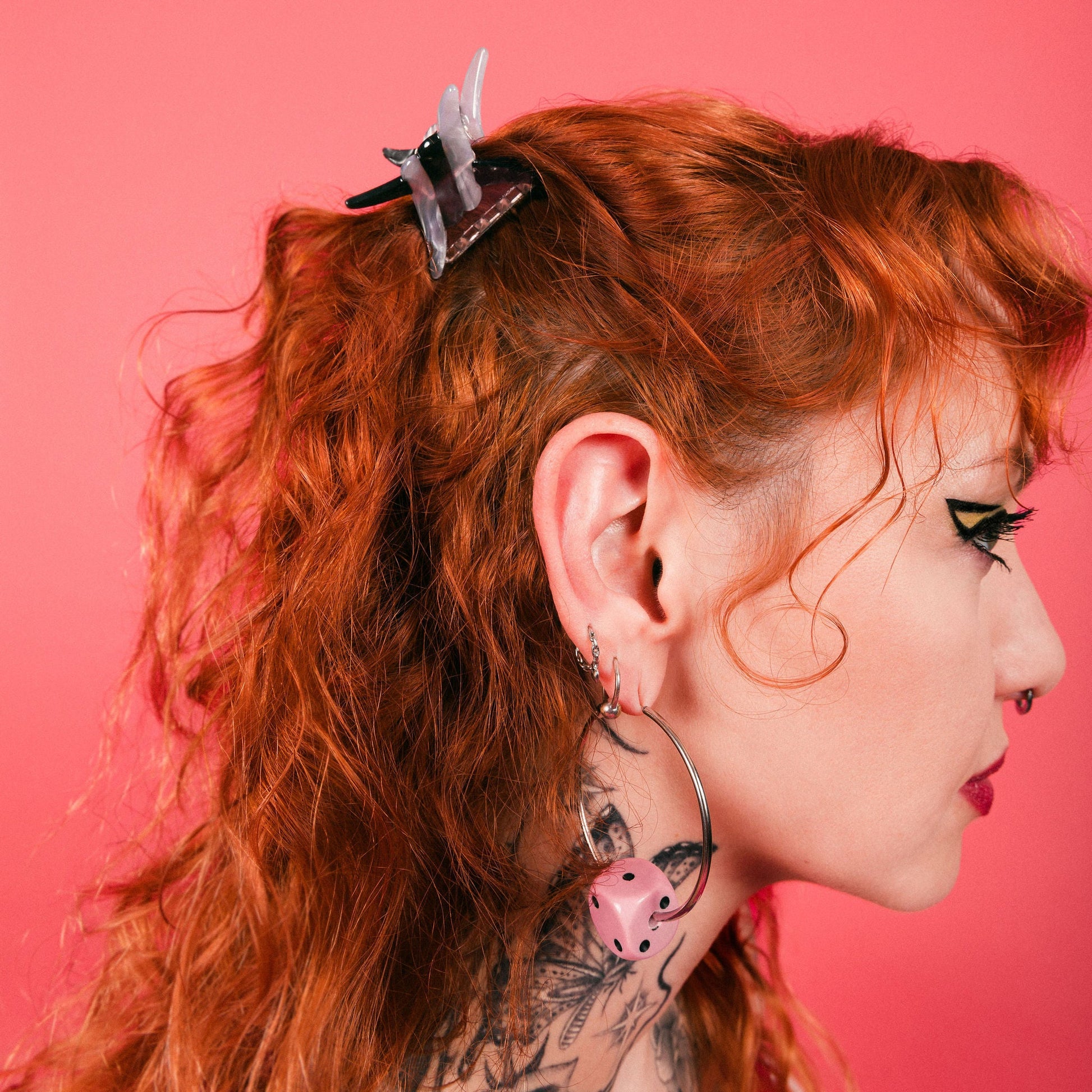 Woman with red hair and tattoos wearing dice hoop earrings and a barbwire mini claw clip pinned back in red hair against a pink backdrop. 