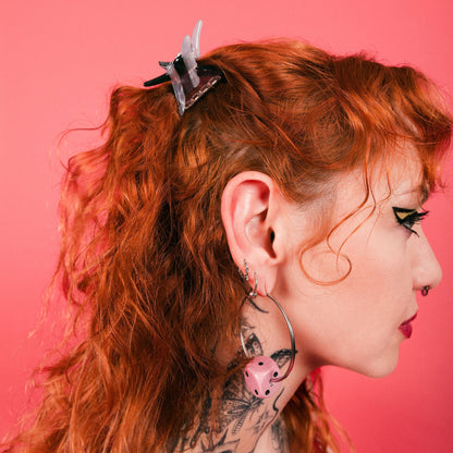 Woman with red hair and tattoos wearing dice hoop earrings and a barbwire mini claw clip pinned back in red hair against a pink backdrop. 