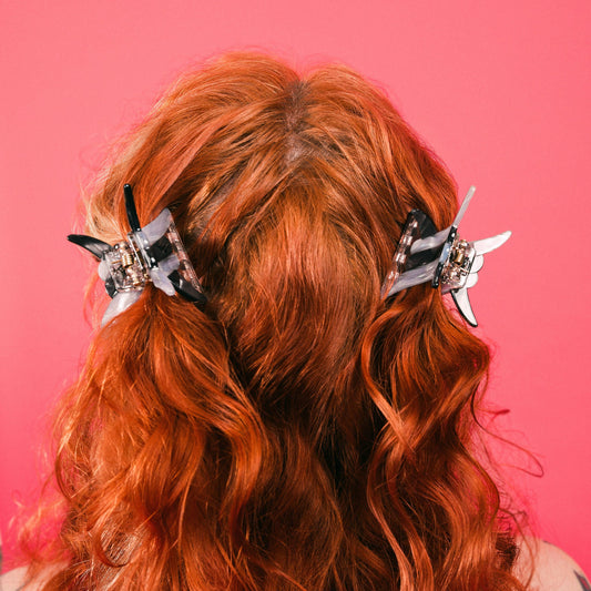 Two mini barbwire claw hair clips clipped onto the back of a head with long red hair against a pink background. 