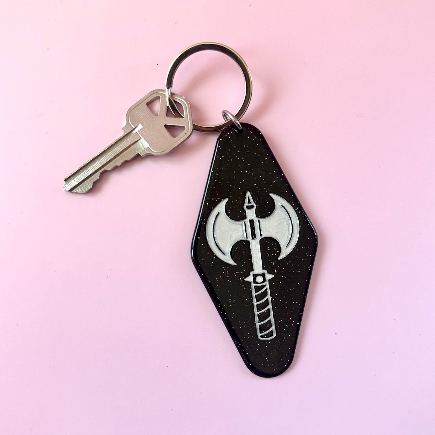 A black coffin-shaped keychain with a silver axe graphic, attached to a metal key ring with a silver key, set against a solid light pink background.