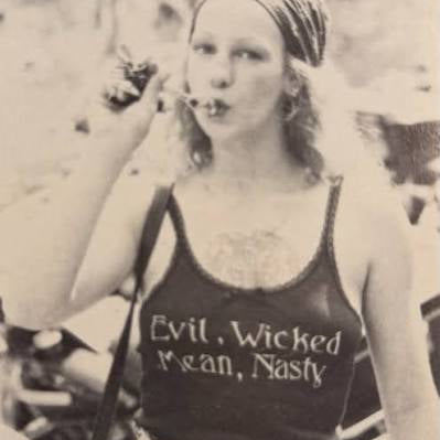 A vintage black-and-white photo of a woman wearing a tank top with the text 'Evil, Wicked Mean, Nasty' printed on the front, paired with jeans and a headscarf.