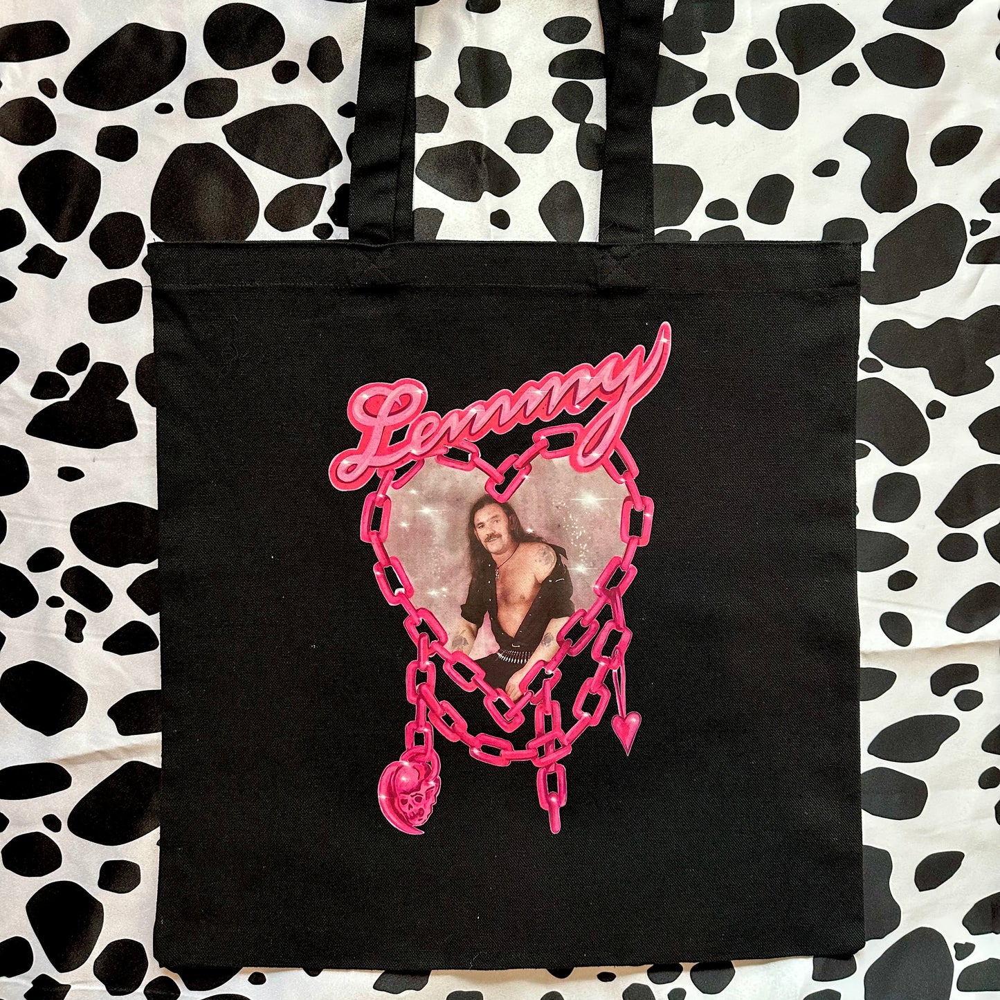 A black canvas tote bag with a pink 'Lemmy' heart graphic, laid flat on a black and white cow print background.