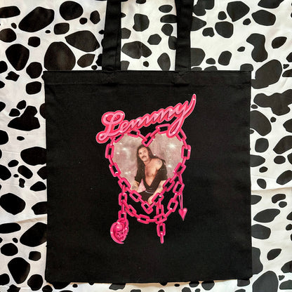 A black canvas tote bag with a pink 'Lemmy' heart graphic, laid flat on a black and white cow print background.