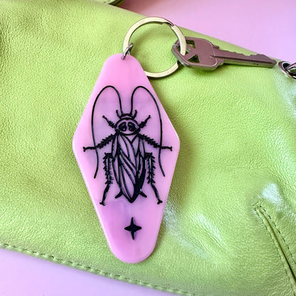 A pink hotel keychain featuring a black line art illustration of a cockroach shown attached to a silver key ring on a light green leather bag against a pink background.