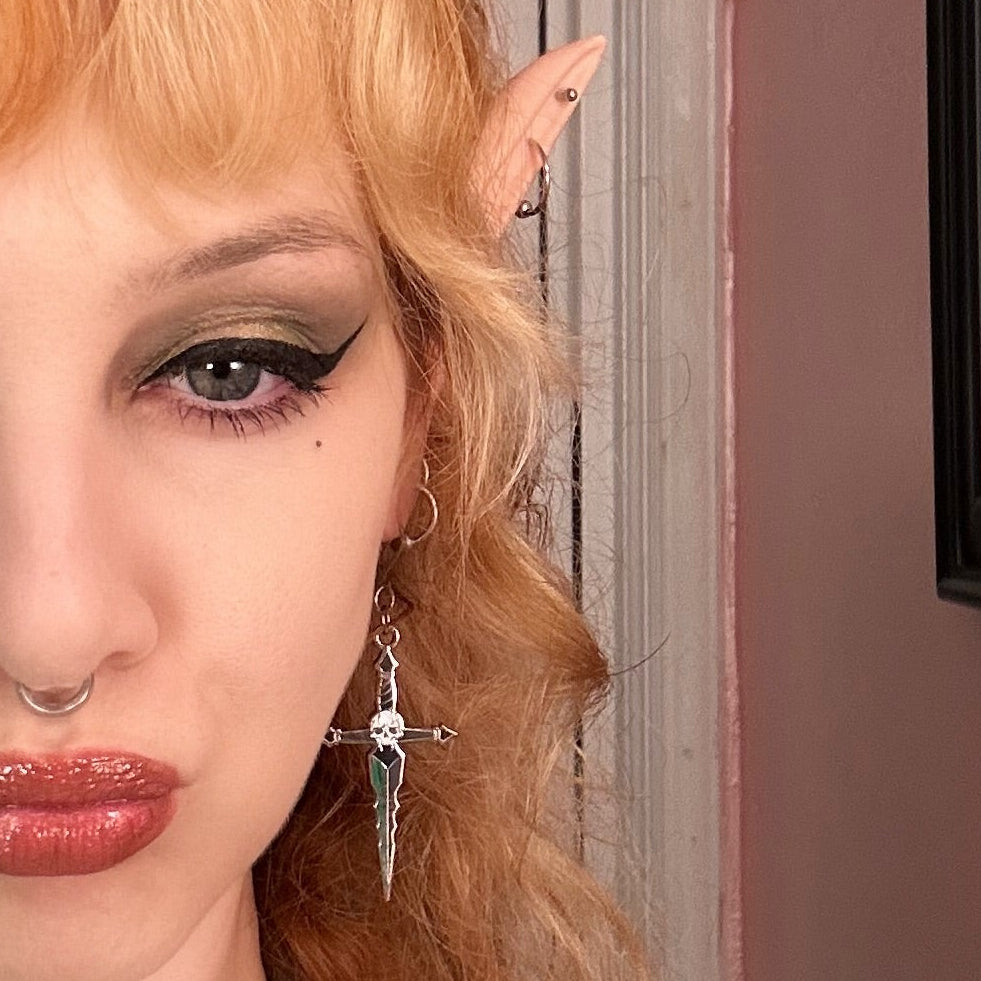 Person with red hair and elf ears in a room with framed pictures on the wall with a punk goth metal enamel skull dagger earring. 