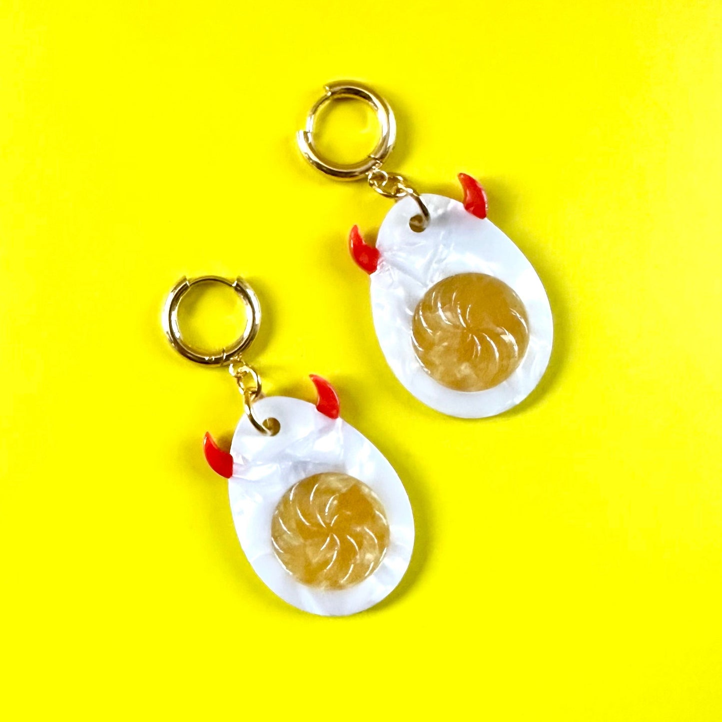 A pair of novelty earrings shaped like hard-boiled eggs with gold-tone metal rings, white bodies, and small red accents, displayed on a bright yellow background.