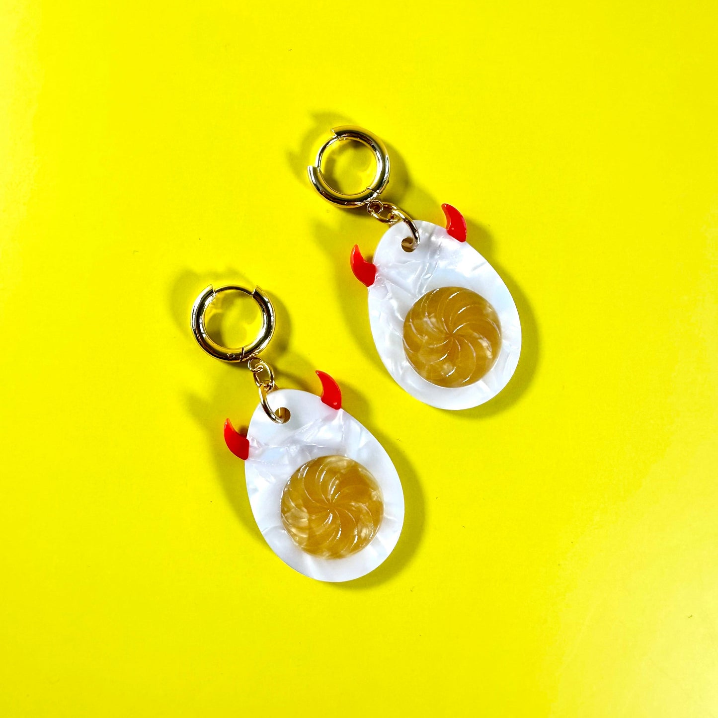 A pair of novelty earrings shaped like fried eggs, featuring white eggshells with gold-colored rims, yellow yolks, and small red accents for the beaks and feet, shown on a bright yellow background.