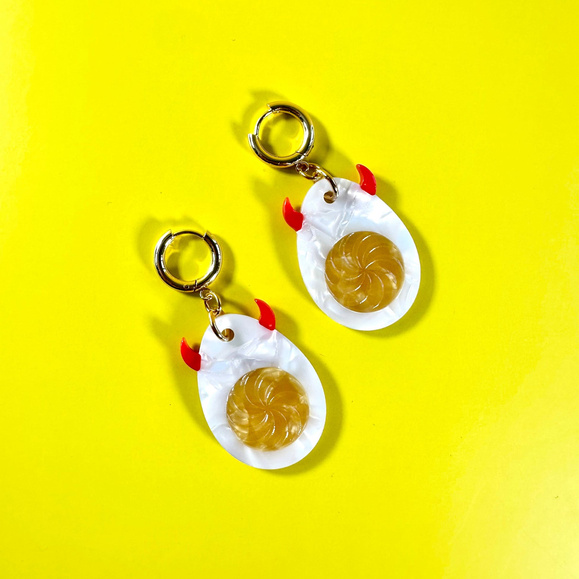 A pair of novelty earrings shaped like fried eggs, featuring white eggshells with gold-colored rims, yellow yolks, and small red accents for the beaks and feet, shown on a bright yellow background.